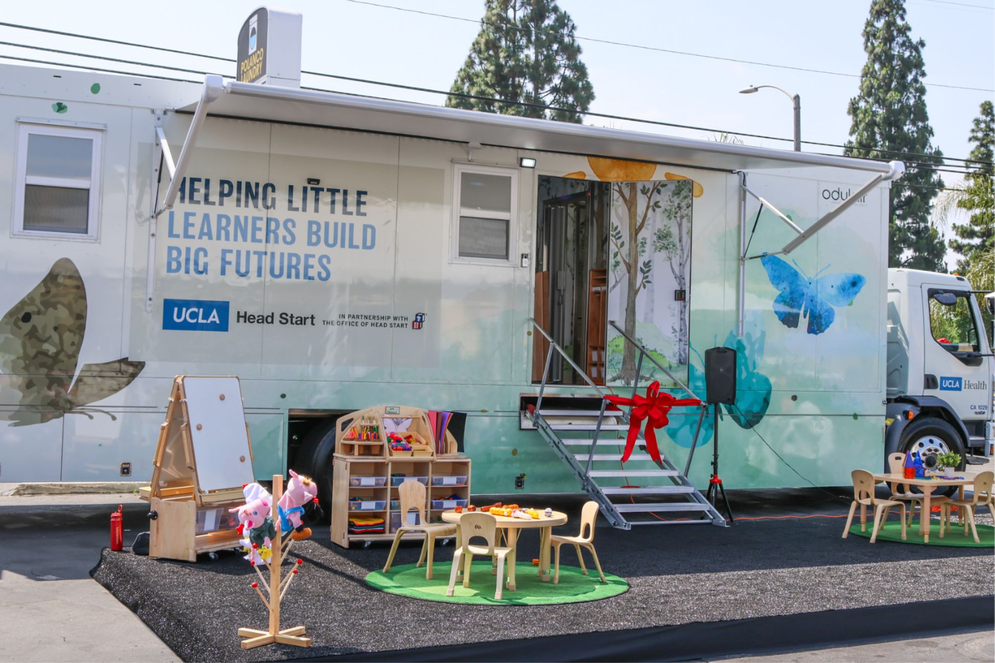 White mobile trailer steps are adorned with tied red ribbon for opening ceremony while gray mat sits underneath two child-size wooden tables small wooden bookcase and white dry-erase board