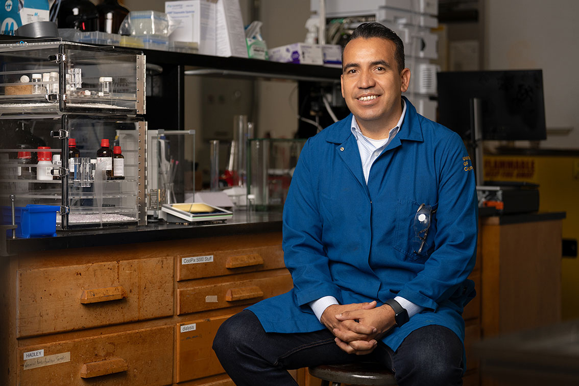 Professor Osvaldo Gutierrez wearing a blue lab coat, sits in front of a lab setting.